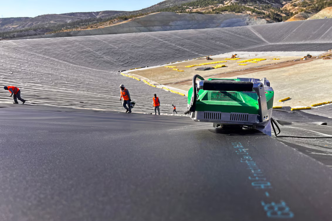 Máquina automática Leister soldando geomembrana em talude de obra de impermeabilização, com equipe ao fundo supervisionando a instalação